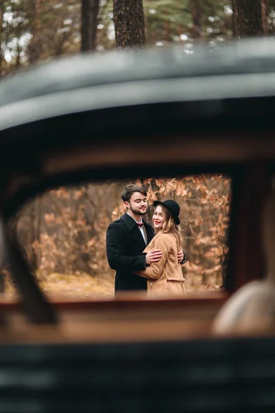 Romantic fairytale wedding couple kissing and embracing in pine forest near retro car.