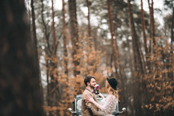 Romantic fairytale wedding couple kissing and embracing in pine forest near retro car.