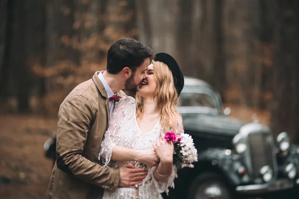 Romantic fairytale wedding couple kissing and embracing in pine forest near retro car.