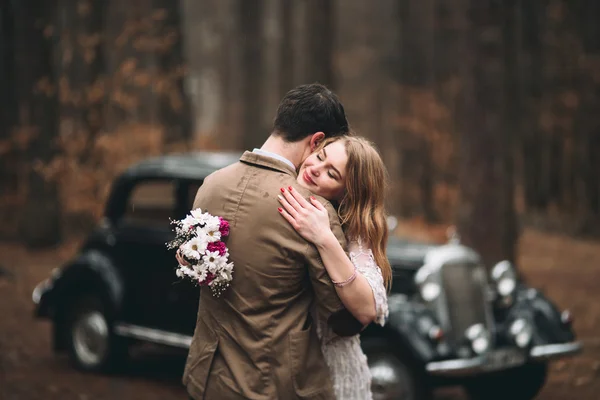 Romantic fairytale wedding couple kissing and embracing in pine forest near retro car.