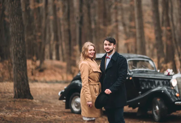 Romantic fairytale wedding couple kissing and embracing in pine forest near retro car.