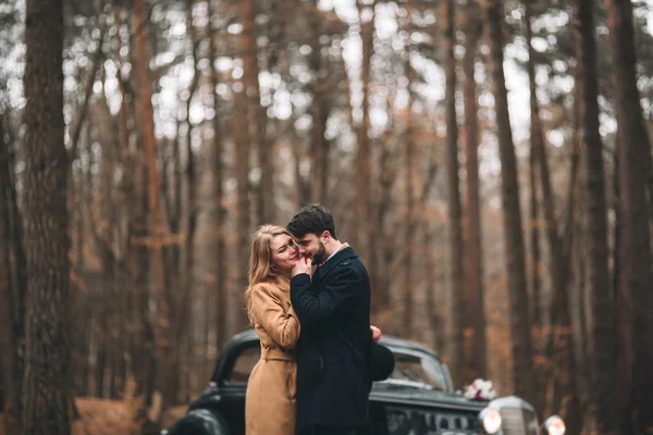 Romantic fairytale wedding couple kissing and embracing in pine forest near retro car.