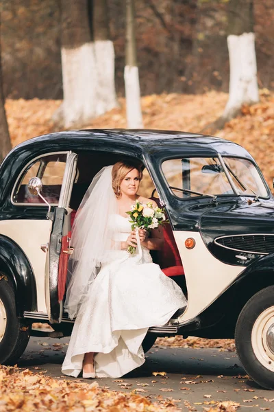 Beautiful happy bride with bouquet near retro car in autumn