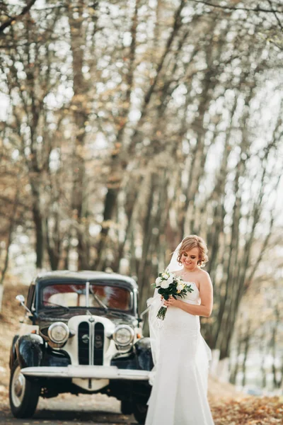 Beautiful happy bride with bouquet near retro car in autumn