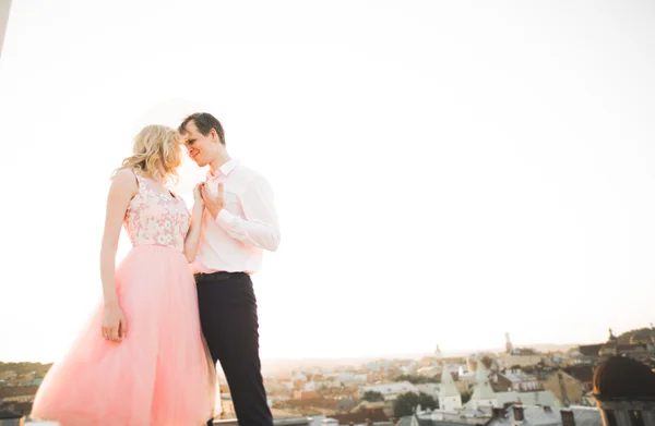 Young couple in love posing on roof with perfect city view holding hands and hugging . Beautiful sunset