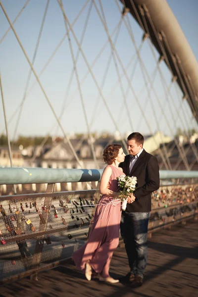 Stylish loving wedding couple, groom, bride with pink dress kissing and hugging on a bridge at sunset