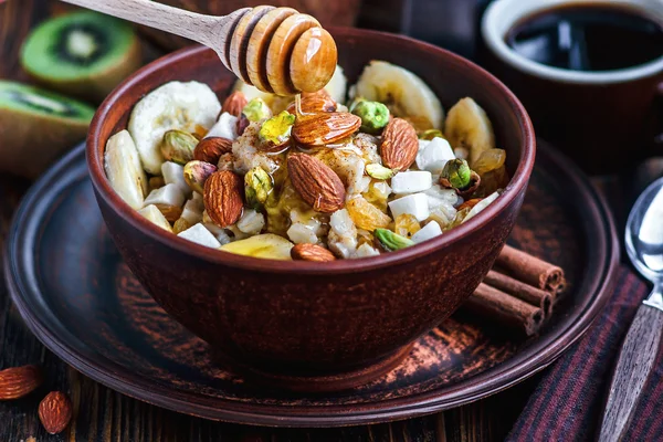 Organic oatmeal porridge with bananas, honey, almonds, pistachio, coconut, kiwi fruit, cinnamon, raisins in dark ceramic bowl and cup of coffee. Healthy breakfast and diet concept on wooden table.