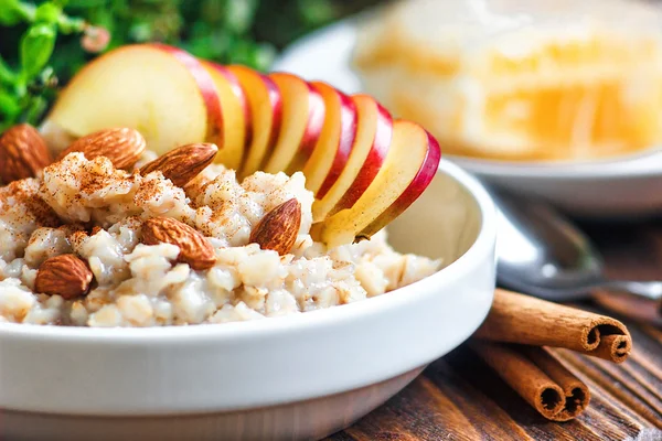 Organic oatmeal porridge in white ceramic bowl with apple, almond, honey and cinnamon. Healthy breakfast - health and diet concept on the wooden table, close up.