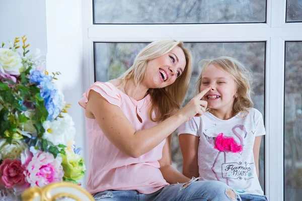 Mom and daughter blonde caucasians are sitting on a windowsill of a large window looking at each other. Beautiful smile. Happy beautiful family. Mothers Day