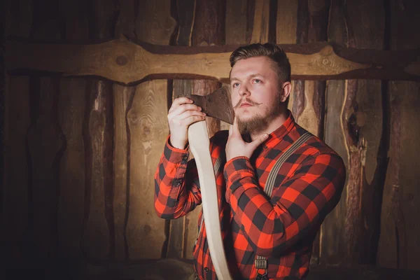 Style lumberjack. Woodcutter with a beard and mustache, wearing a red shirt, pants and suspenders with an ax on a wooden background. Modern style. Thoughtful lumberjack