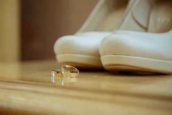 Two gold wedding rings on the heels of white wedding bridal shoes on a wooden table