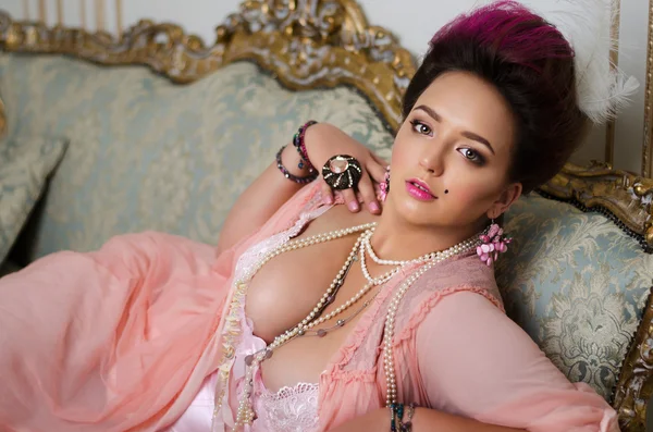 Beautiful brunette with curvaceous, Rococo hair, rings and beads lies on a beautiful sofa in pink peignoir