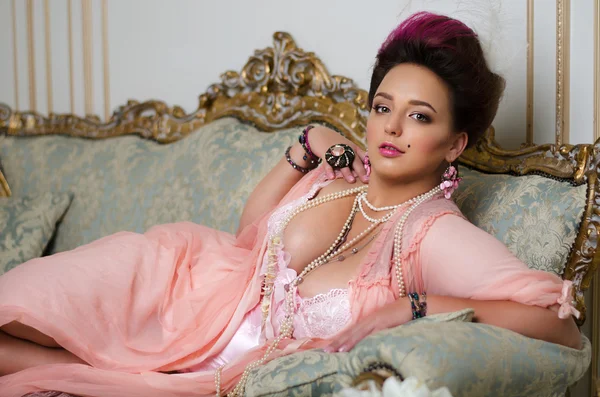 Beautiful brunette with curvaceous, Rococo hair, rings and beads lies on a beautiful sofa in pink peignoir