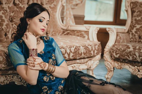 Beautiful woman in a traditional Chinese outfit Indian blue, sitting on the floor in a luxury sofa. On her hands painted with henna mehendi