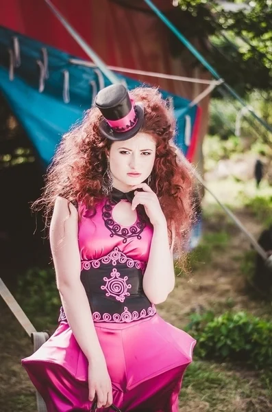 Beautiful girl with red hair in a funny designer pink dress and hat on his head, is behind the scenes of the circus. Clown. Circus tent