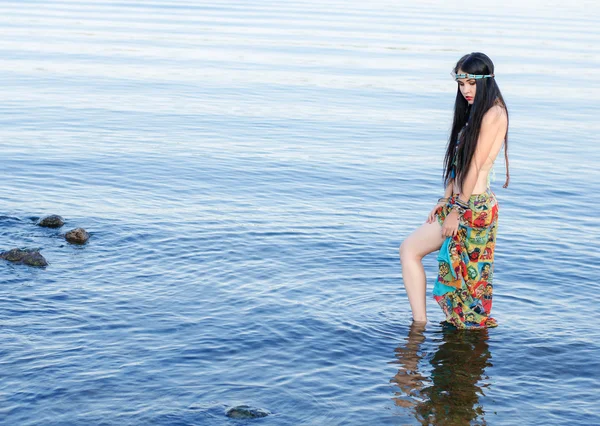 Beautiful slim woman with long black hair and a bandage on his head, in the clothes in boho style, standing in water.  Boho chic style