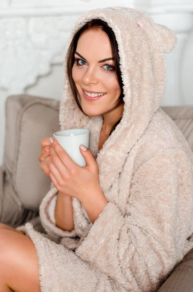 Beautiful young brunette woman in a home beige terry robe with hood sitting on an easy chair at home and holding a mug of coffee, tea and smiling