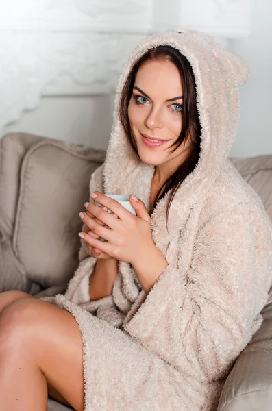 Beautiful young brunette woman in a home beige terry robe with hood sitting on an easy chair at home and holding a mug of coffee, tea and smiling