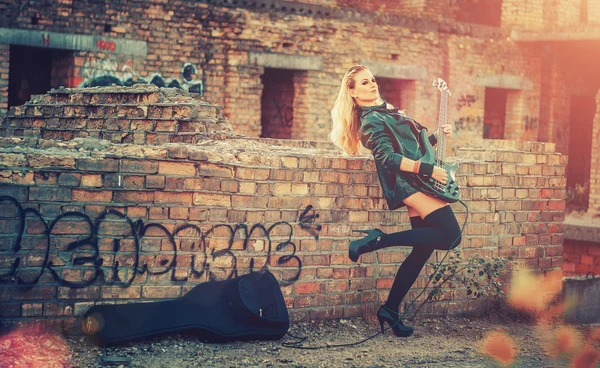 Beautiful young blond woman in black body suit, black leather jacket and black shoes and socks, stands on the roof of an abandoned nedostorennogo home, with brick walls and playing on black guitar