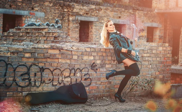Beautiful young blond woman in black body suit, black leather jacket and black shoes and socks, stands on the roof of an abandoned nedostorennogo home, with brick walls and playing on black guitar