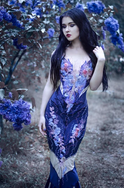 Pretty slim girl with dark hair in a long emerald green dress with bare shoulders is in the garden with blooming lilac bushes