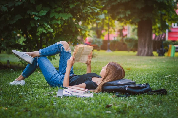 Girl student in a park lying on the grass and reading a book, lying next to her backpack and a stack of notebooks. Education concept