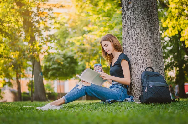 Happy girl student sitting on the grass near the tree in the park and reading. Education concept