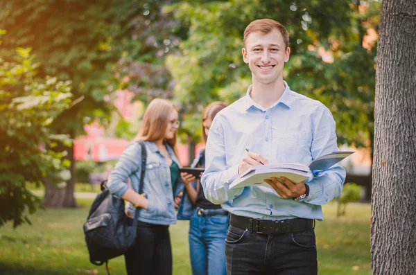 Happy smiling boy student in braces, shirt and trousers with a book in the summer park, behind it there are girls and communicate