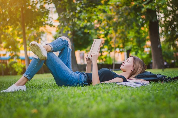 Girl student in a park lying on the grass and reading a book, lying next to her backpack and a stack of notebooks. Education concept