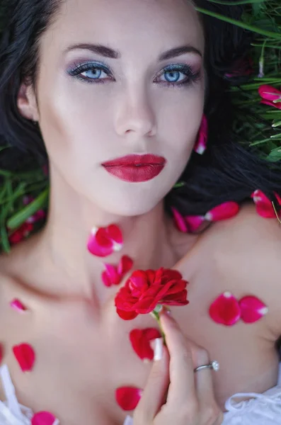 Portrait of a beautiful young brunette with red lips, blue eyes, lying on green grass with rose petals