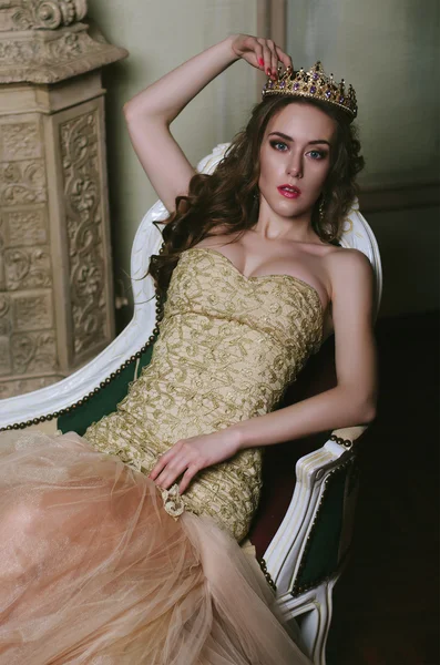 Beautiful brown-haired woman with magnificent breasts in a white dress and a crown on her head lying on beautiful sofa