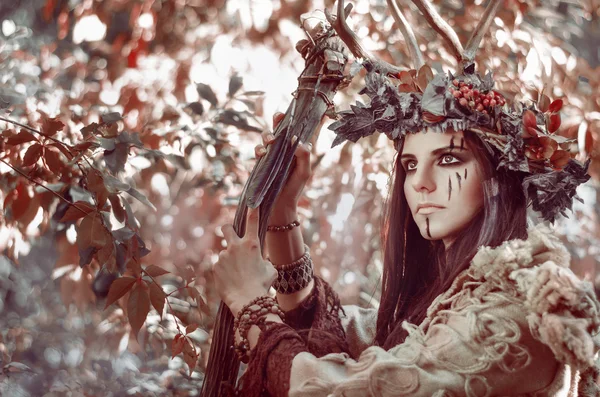 Portrait of a beautiful brunette with a painted face, a floral wreath on her head and horns in the form of shaman, with the village staff in hand