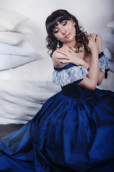 Beautiful young brunette in blue historic, vintage dress sitting on the floor next to a vintage bed