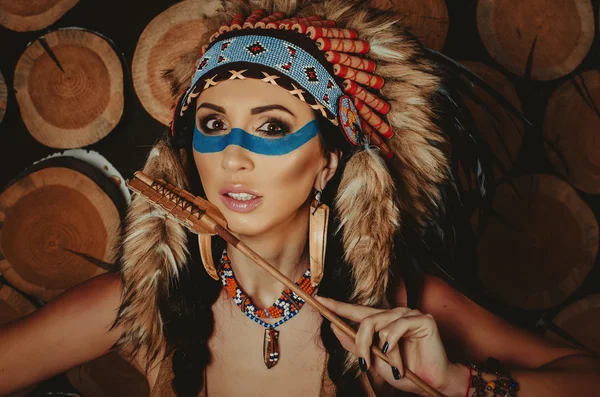 Beautiful girl with painted face in the national Indian suit with ethnic ornaments, earrings in the form of fangs, on a background of wooden logs