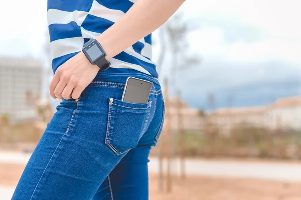 Back side view of woman in jeans using smart watch with mobile phone in pocket. Close up.