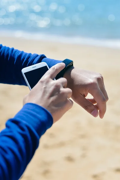 Person holding phone checking connection using smart watch on the sandy beach. Sunny background outdoors