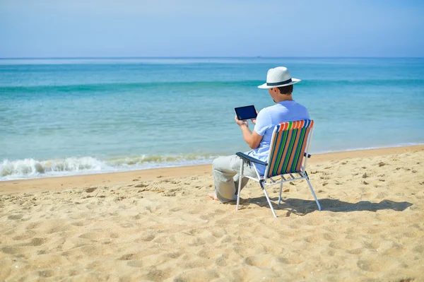 Man using tablet relaxing on ocean beach, blue sky outdoors background