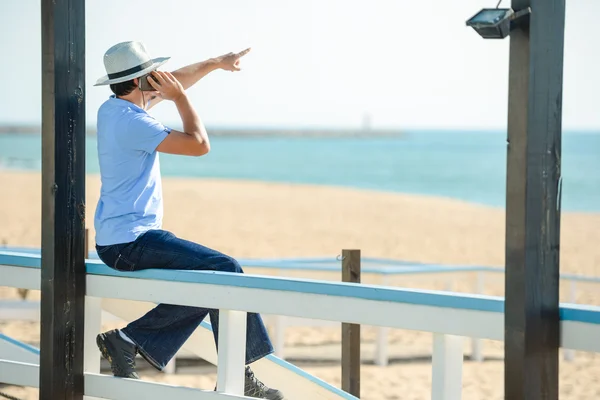 Male wear hat talking on phone and pointing at sea and blue sky.