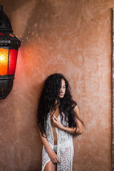 Beautiful woman with long black hair in white lace lingerie in room with vintage lamp