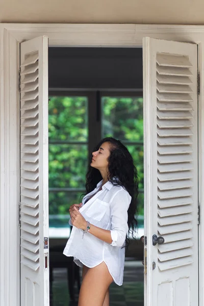 Beautiful woman with long black hair posing near white door in black lingerie. Morning mood