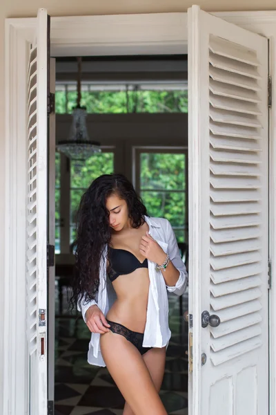 Beautiful woman with long black hair posing near white door in black lingerie. Morning mood
