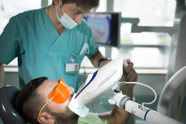 Man having teeth whitening by dental UV whitening device,dental assistant taking care of patient,eyes protected with glasses.