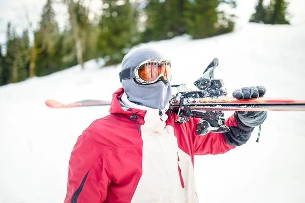 Young man holding ski at ski resort.Skier holding skis looking at the mountains.Side view of handsome skier man with mask and holding ski equipment