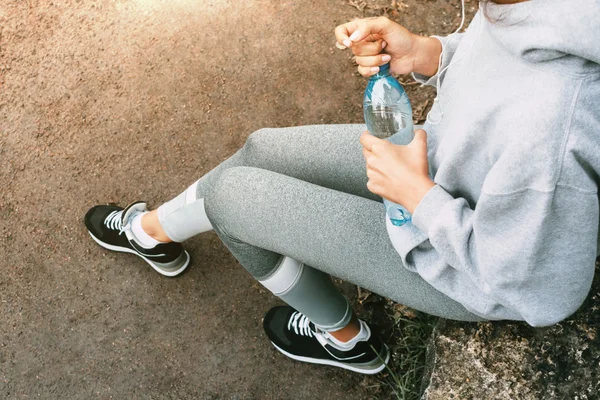 Woman in sportswear sitting in the park with a bottle of water and resting after a workout