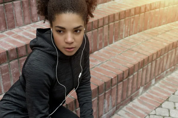 Young sporty African American woman wearing earphones in urban environment preparing for workout