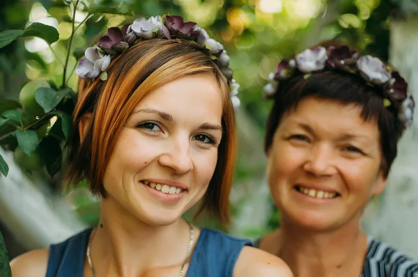 Portrait of a smiling mother and daughter are posing in wreaths of artificial flowers from the outdoors on a background of trees. The concept of family. The relationship of the mother and daughter.