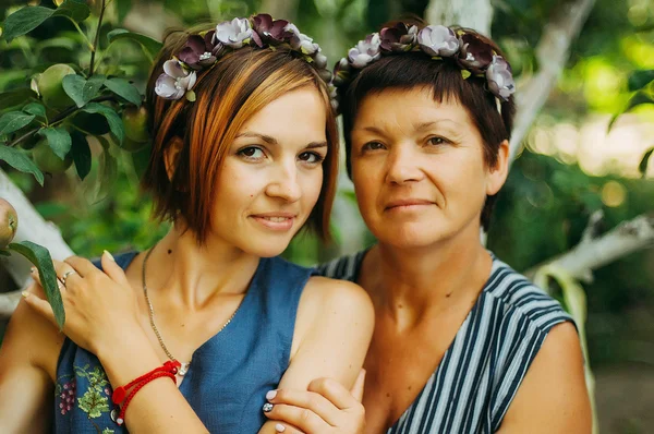 Portrait of a smiling mother and daughter are posing in wreaths of artificial flowers from the outdoors on a background of trees. The concept of family. The relationship of the mother and daughter.