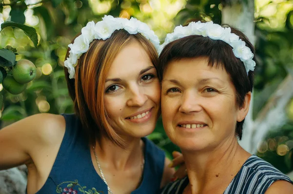 Portrait of a smiling mother and daughter are posing in wreaths of artificial flowers from the outdoors on a background of trees. The concept of family. The relationship of the mother and daughter.