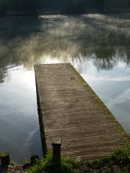 Wooden jetty on the River Thames with mist floating above the river and reflections of vapour trails on the glassy surface of the water