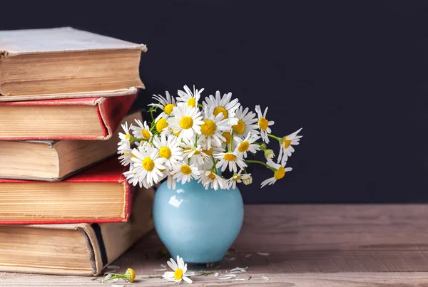 A stack of old vintage books lying on a wooden shelf with a bouquet of daisies in a blue vase. Country still life.